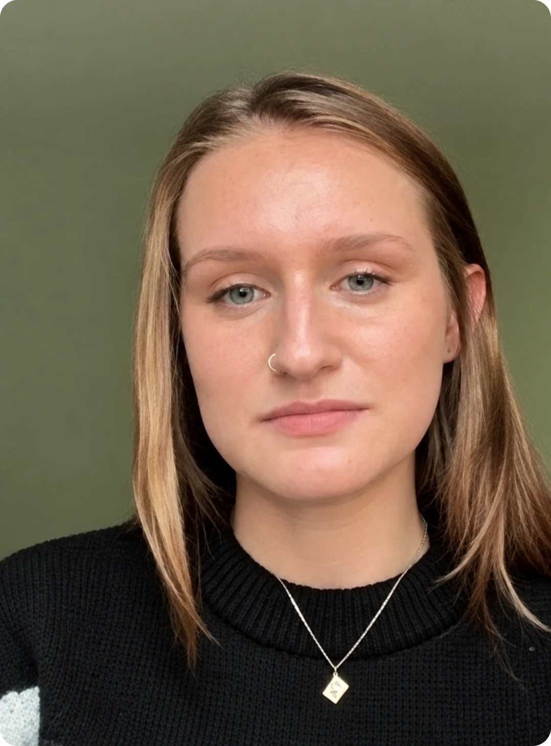 Portrait of a young woman with light brown hair wearing a black sweater and a necklace with a square pendant.