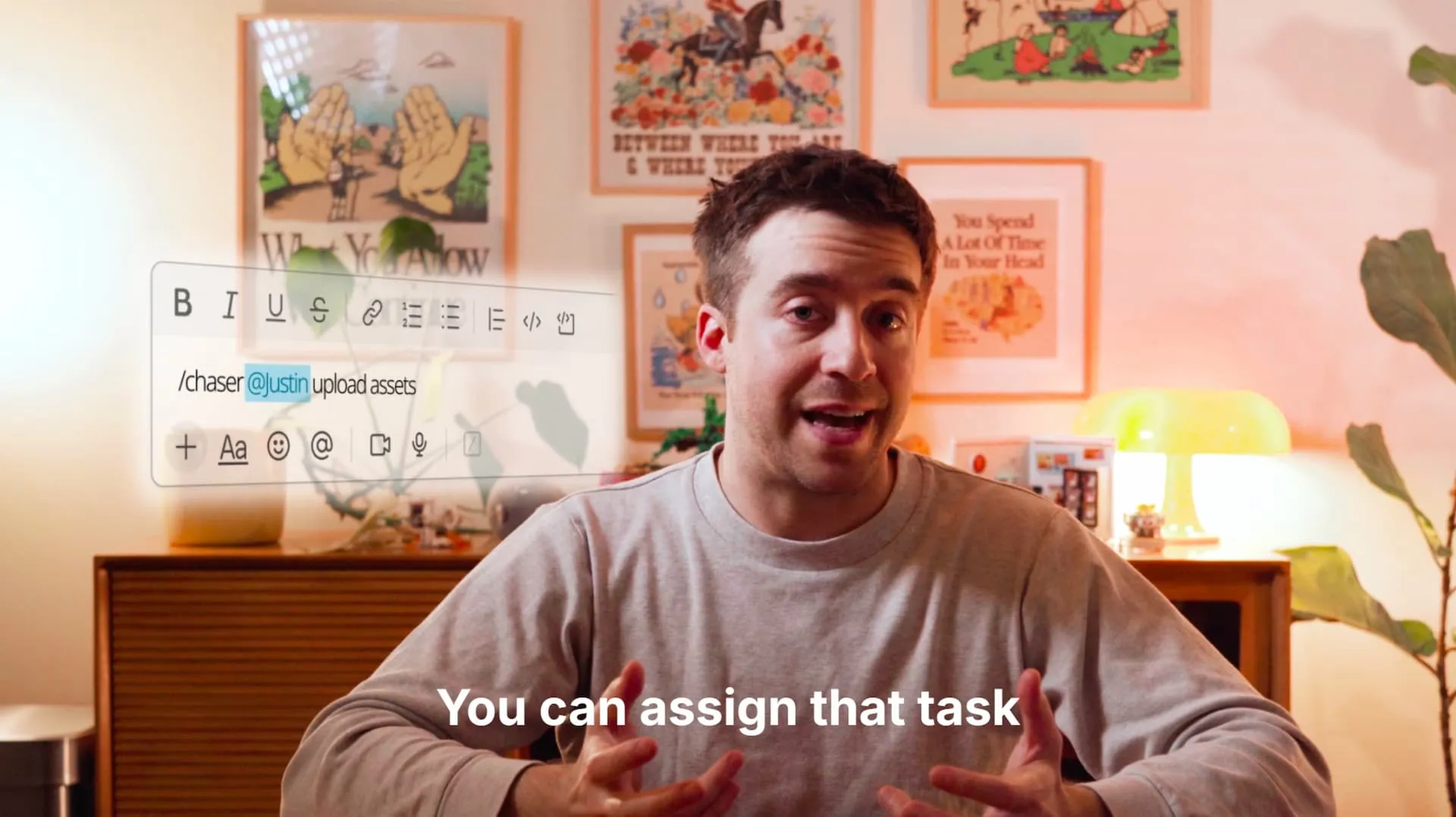 Man in gray sweatshirt explaining task assignment with text overlay 'You can assign that task' and a digital interface showing a message tagging '@Justin' to upload assets.